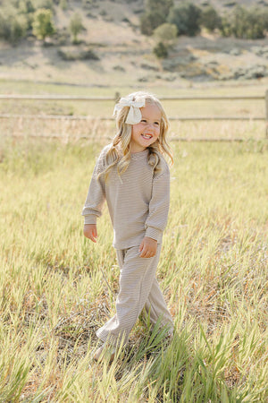 A smiling child wears a striped loungewear cotton set, featuring a long-sleeve top and pants, while standing in a grassy field with a rustic wooden fence in the background.