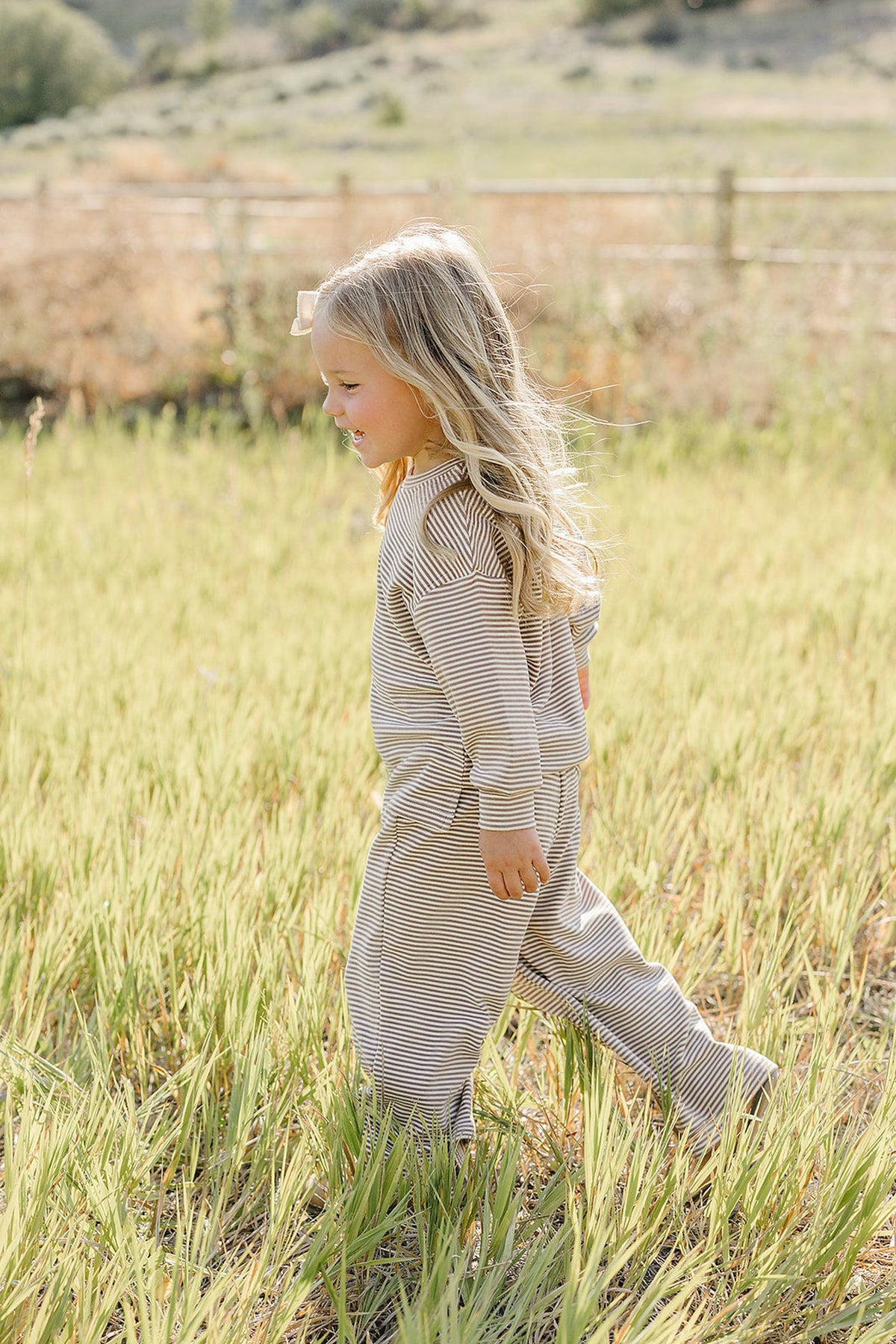 A young child with long, wavy hair joyfully runs through a grassy field, wearing a soft, striped loungewear set in neutral tones, perfect for comfort and play.