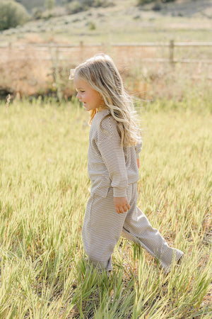 A young child with long, wavy hair joyfully runs through a grassy field, wearing a soft, striped loungewear set in neutral tones, perfect for comfort and play.