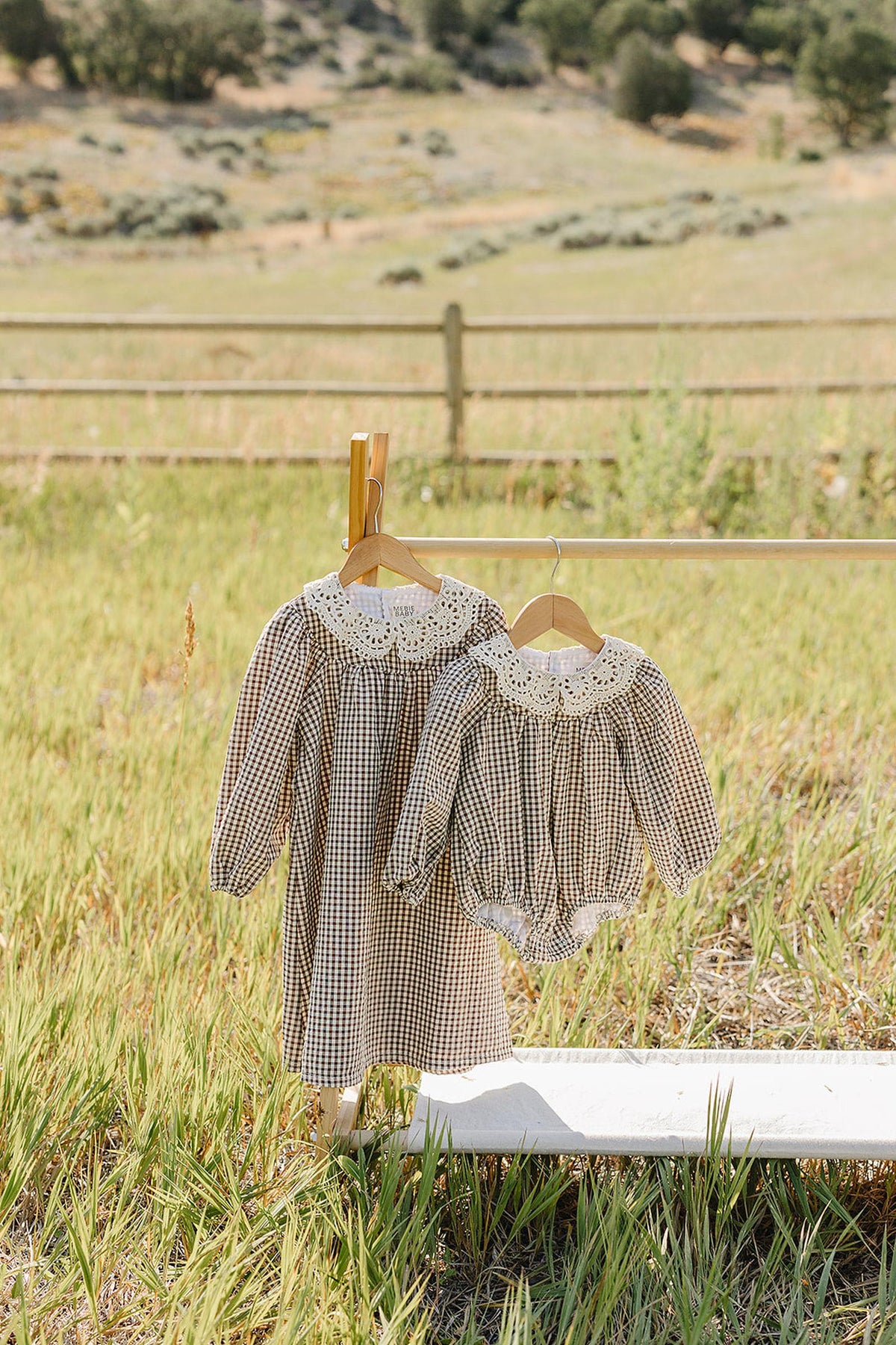 A charming camel gingham lace bubble cotton romper and matching dress hang on a wooden hanger against a serene, grassy landscape, showcasing their soft, breathable fabric and delicate lace detailing.