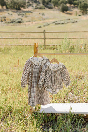 A charming camel gingham lace bubble cotton romper and matching dress hang on a wooden hanger against a serene, grassy landscape, showcasing their soft, breathable fabric and delicate lace detailing.