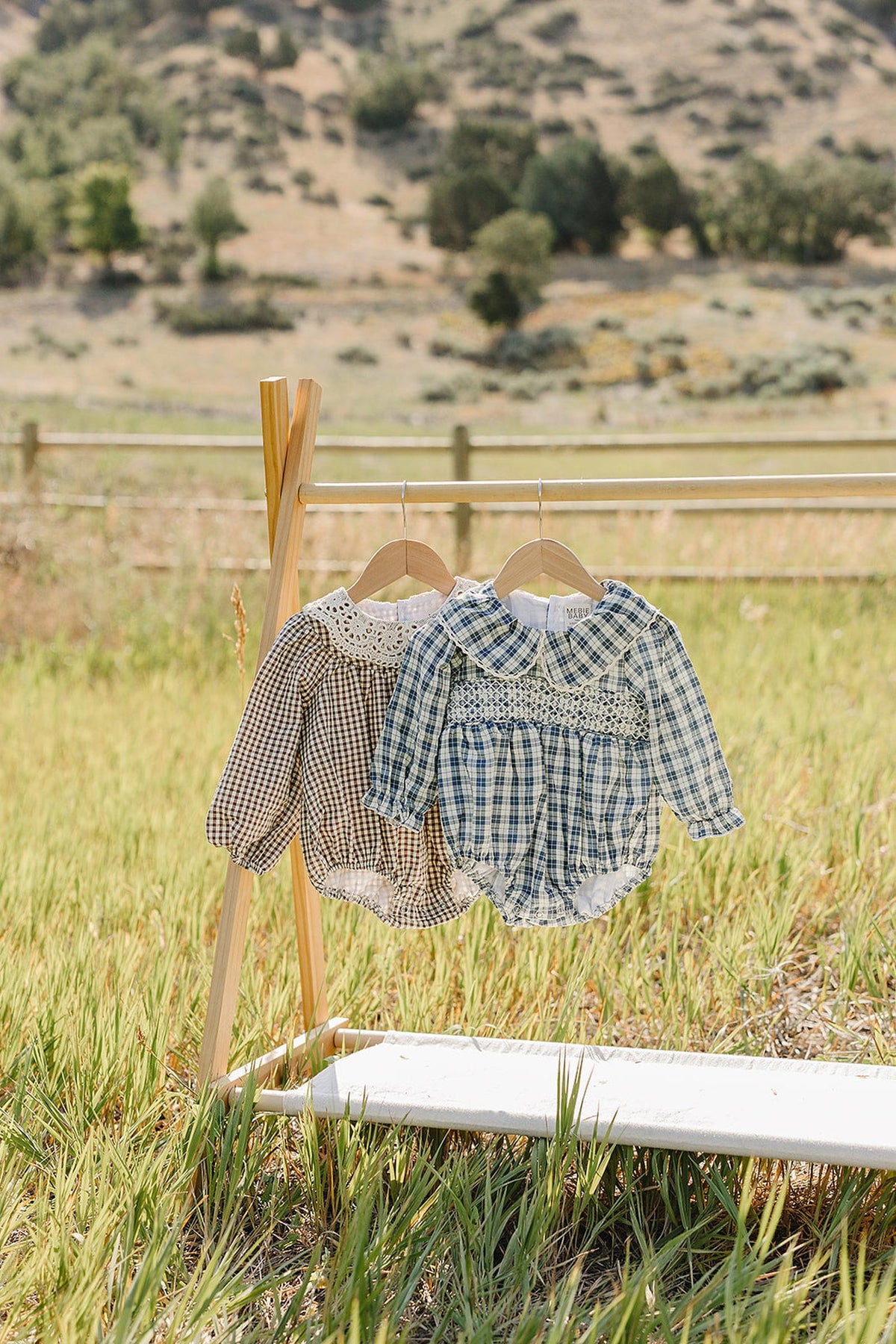 Two bubble rompers hang on a wooden rack in a grassy field, featuring a brown checkered pattern and a blue plaid design, both with ruffle collars and soft cotton fabric.