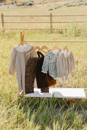 A collection of childrens clothing hangs on a wooden rack in a grassy field, featuring stylish patterns like gingham and leopard print, showcasing soft, comfortable fabrics ideal for everyday wear.