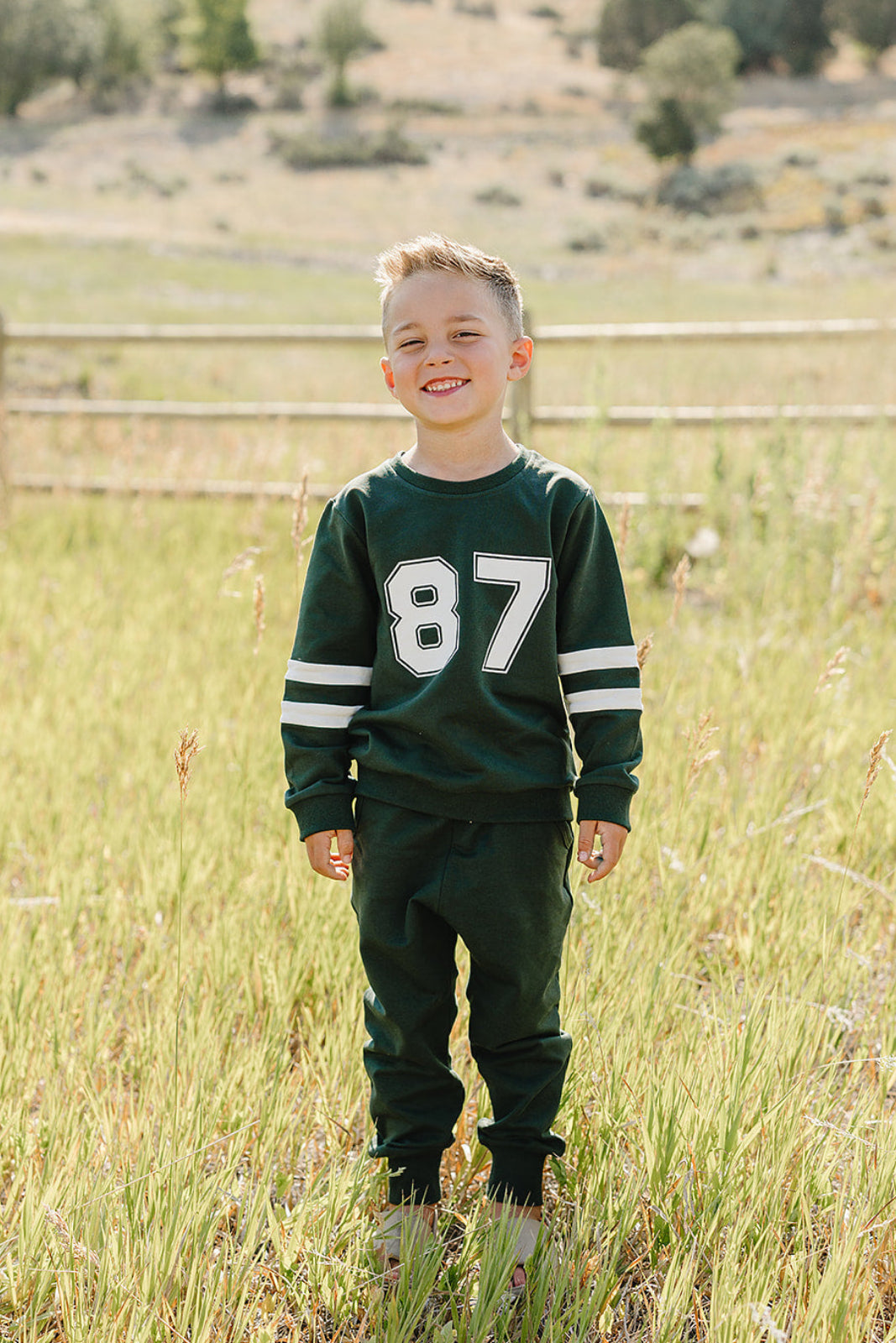 A young child stands smiling in a grassy field, wearing a dark green French terry cotton set with white stripes and the number 87 on the shirt.