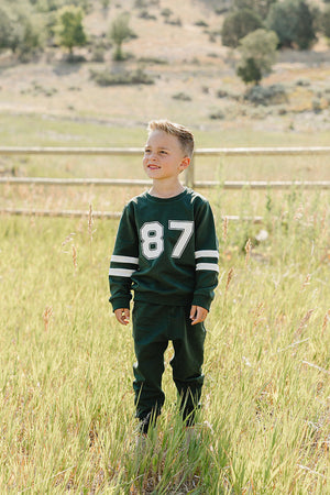 A young boy stands in a grassy field, wearing a dark green French terry cotton set featuring a sporty design with the number 87 and white stripes on the sleeves.