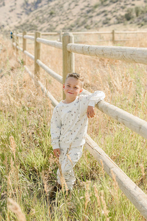 A young boy in a light beige, patterned French terry cotton set leans against a wooden fence in a grassy field, enjoying a sunny day outdoors.