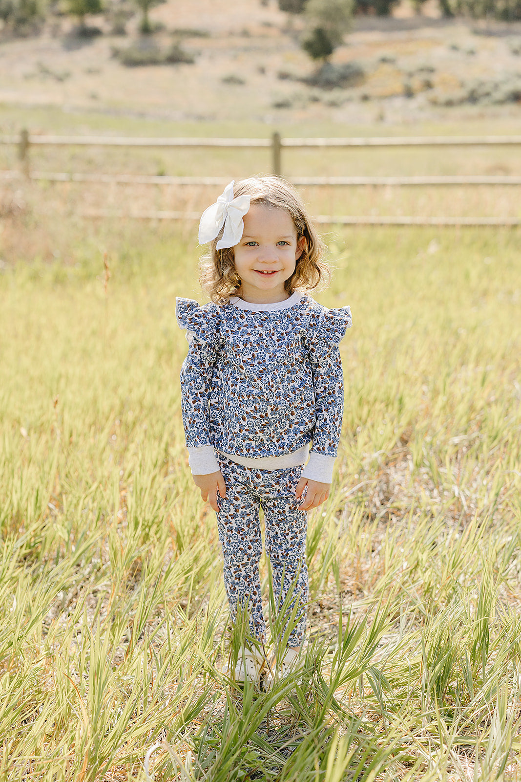 A young girl stands in a grassy field wearing a blue floral ruffle long-sleeve cotton set with matching pants. She has a white bow in her hair, smiling brightly.