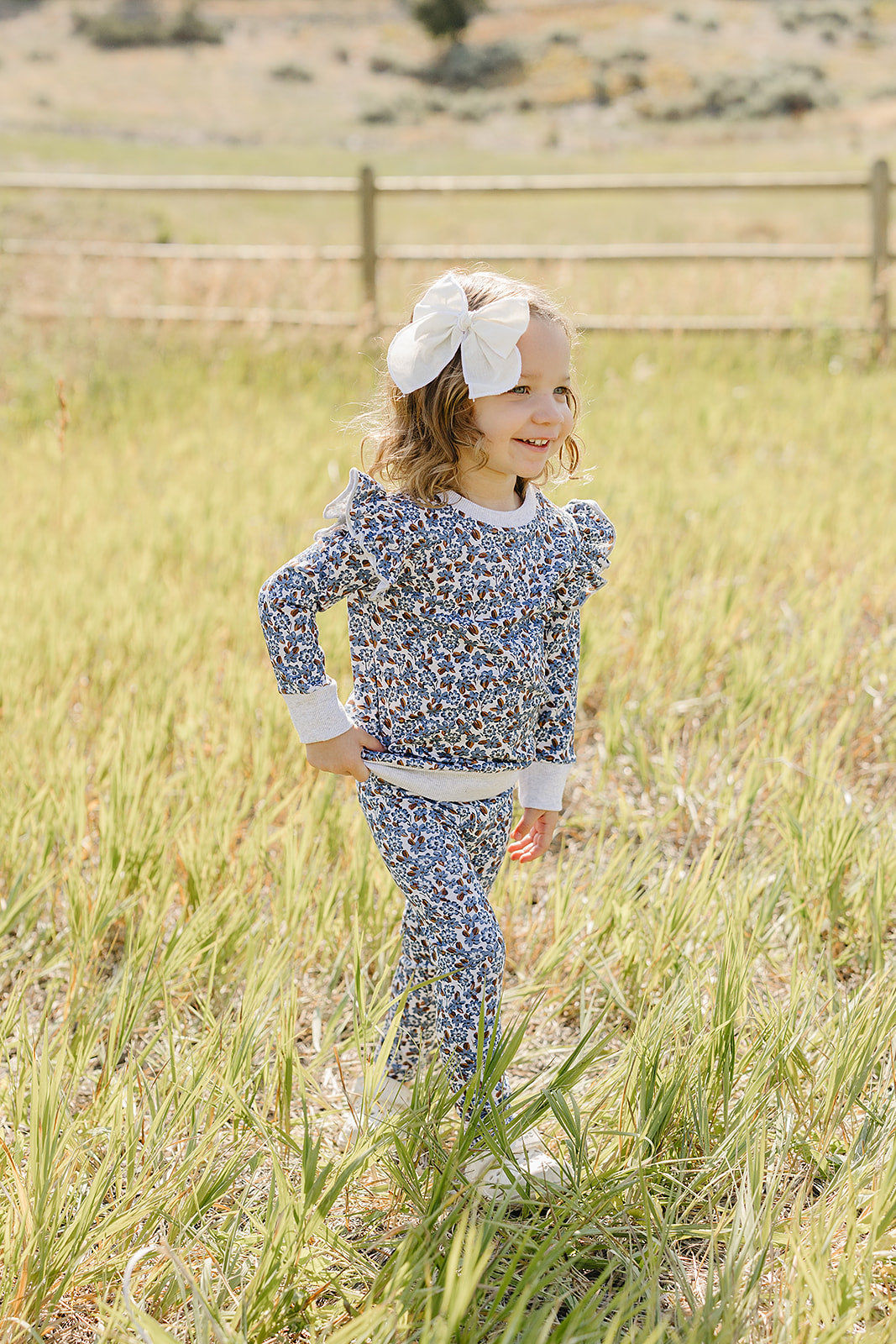 A smiling child wears a floral print long-sleeve top and matching pants in a grassy field. The outfit features ruffle details and is made from soft, breathable fabric, perfect for.