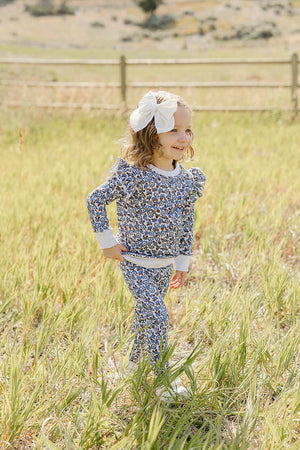 A smiling child wears a floral print long-sleeve top and matching pants in a grassy field. The outfit features ruffle details and is made from soft, breathable fabric, perfect for.