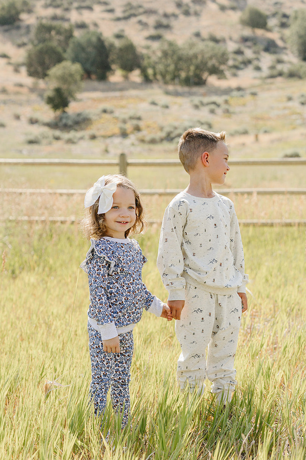 Two children stand in a grassy field, holding hands. The girl wears a blue floral long sleeve top with a white bow, while the boy sports a light-colored outfit with.