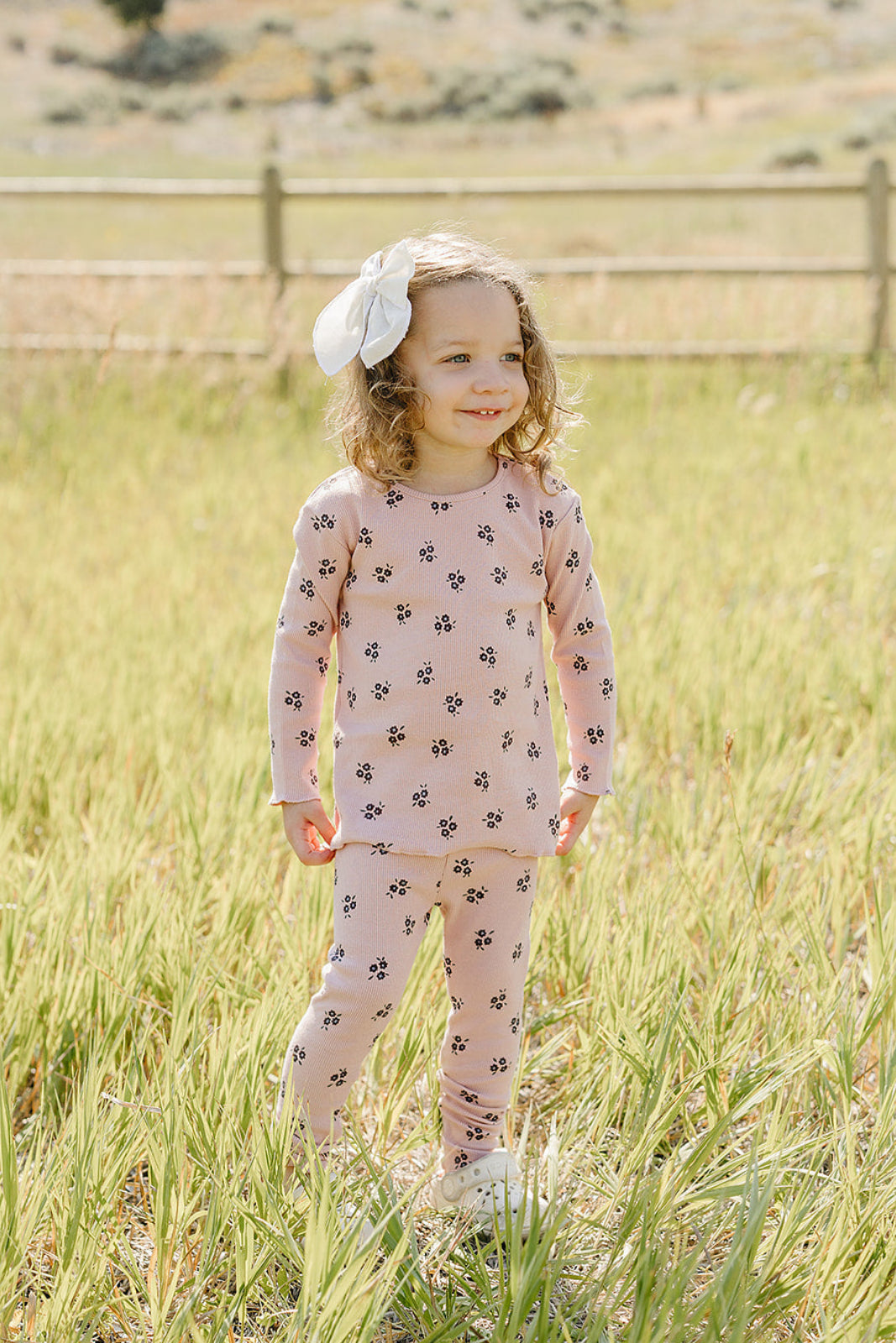 A young child stands in a grassy field, wearing a soft pink cotton outfit decorated with black floral patterns. She smiles and sports a white bow in her curly hair.