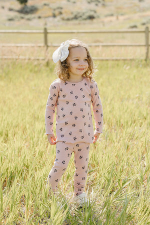 A young child stands in a grassy field, wearing a soft pink cotton outfit decorated with black floral patterns. She smiles and sports a white bow in her curly hair.