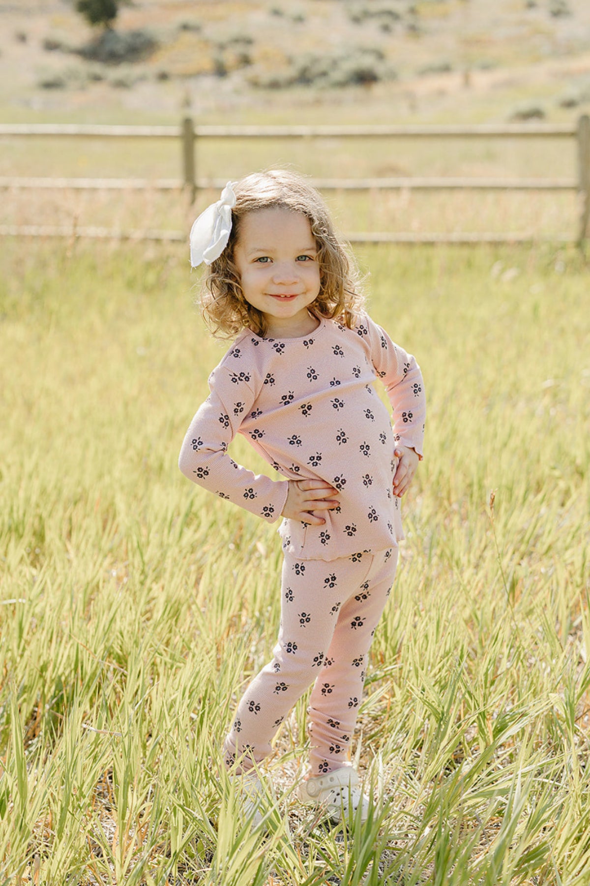 A smiling young child stands in a grassy field, wearing a pink cotton outfit adorned with floral patterns.