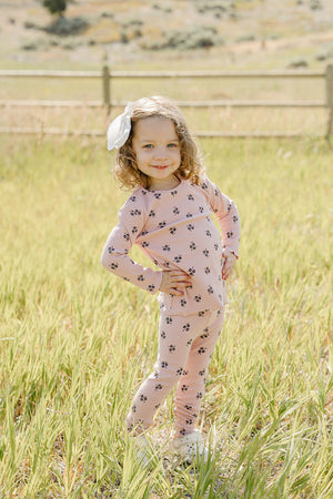 A smiling young child stands in a grassy field, wearing a pink cotton outfit adorned with floral patterns.