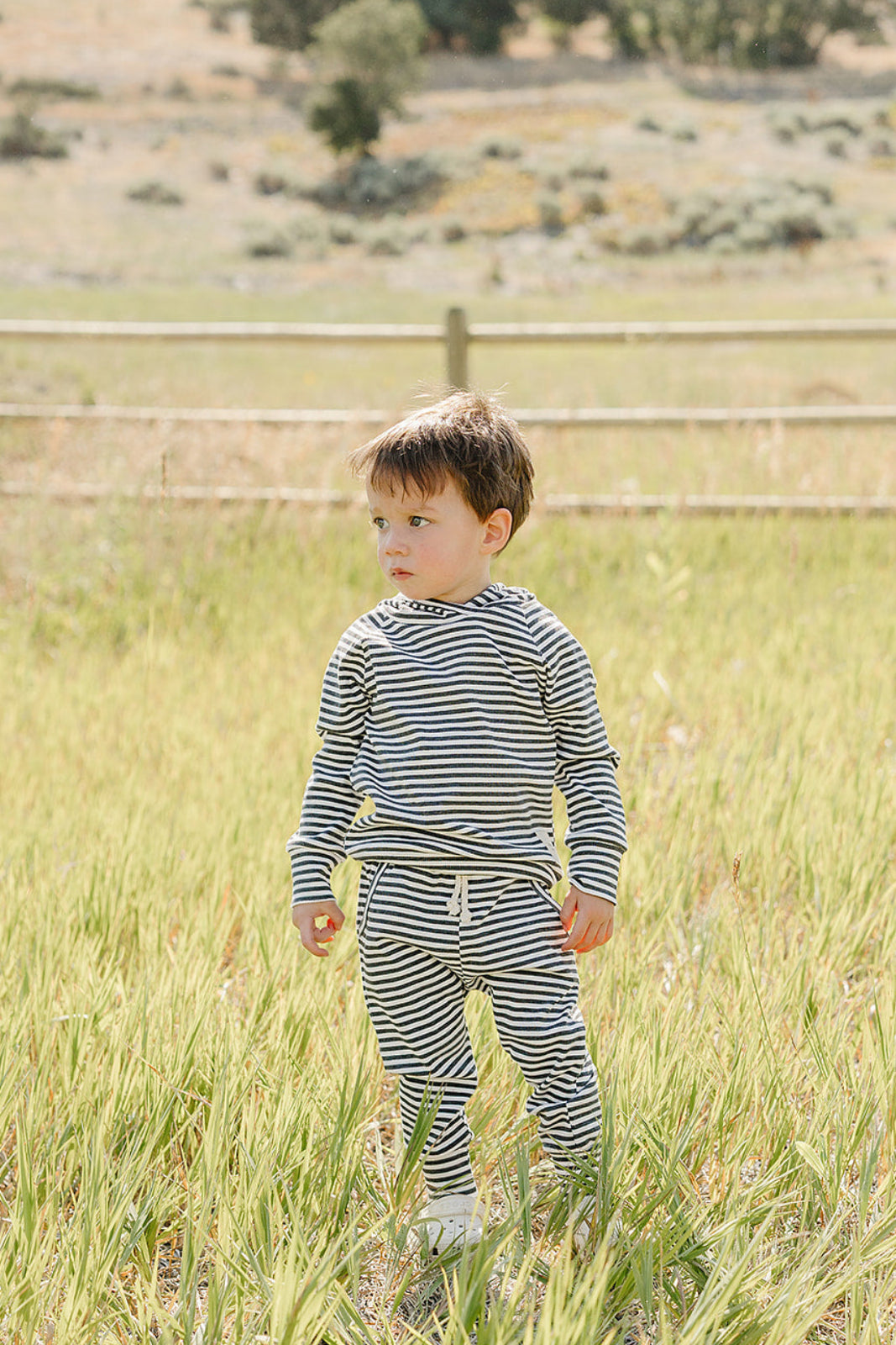 A young child stands in a grassy field, wearing a charcoal stripe ribbed hooded set. The outfit features soft, breathable fabric, perfect for outdoor play and comfort.