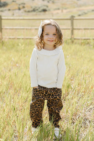 A young child stands in a grassy field wearing a white ribbed top and stylish cheetah-print barrel jeans. The outfit is playful and perfect for outdoor adventures.