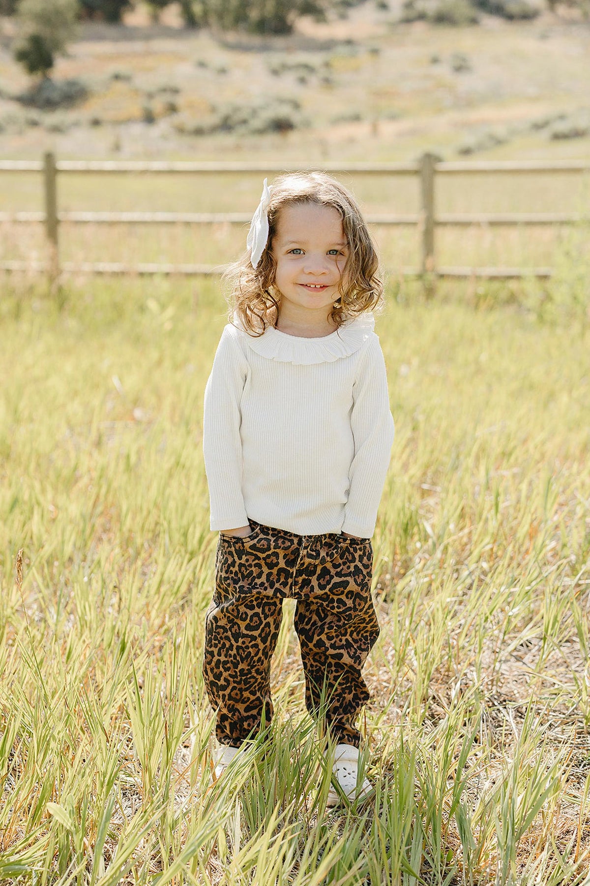 A smiling child stands in a grassy field, wearing a white ribbed long-sleeve top with a ruffle collar and stylish leopard print pants.