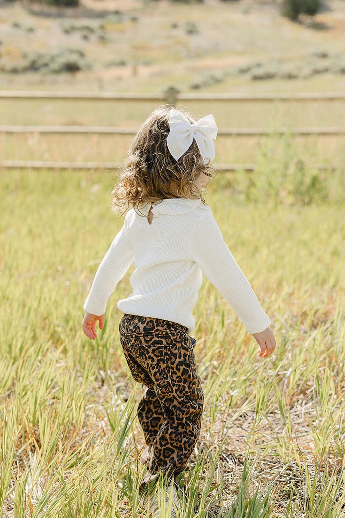 A child with curly hair and a large white bow walks through a grassy field, wearing stylish cheetah print pants and a white sweater, enjoying a sunny outdoor setting.