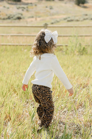 A child with curly hair and a large white bow walks through a grassy field, wearing stylish cheetah print pants and a white sweater, enjoying a sunny outdoor setting.