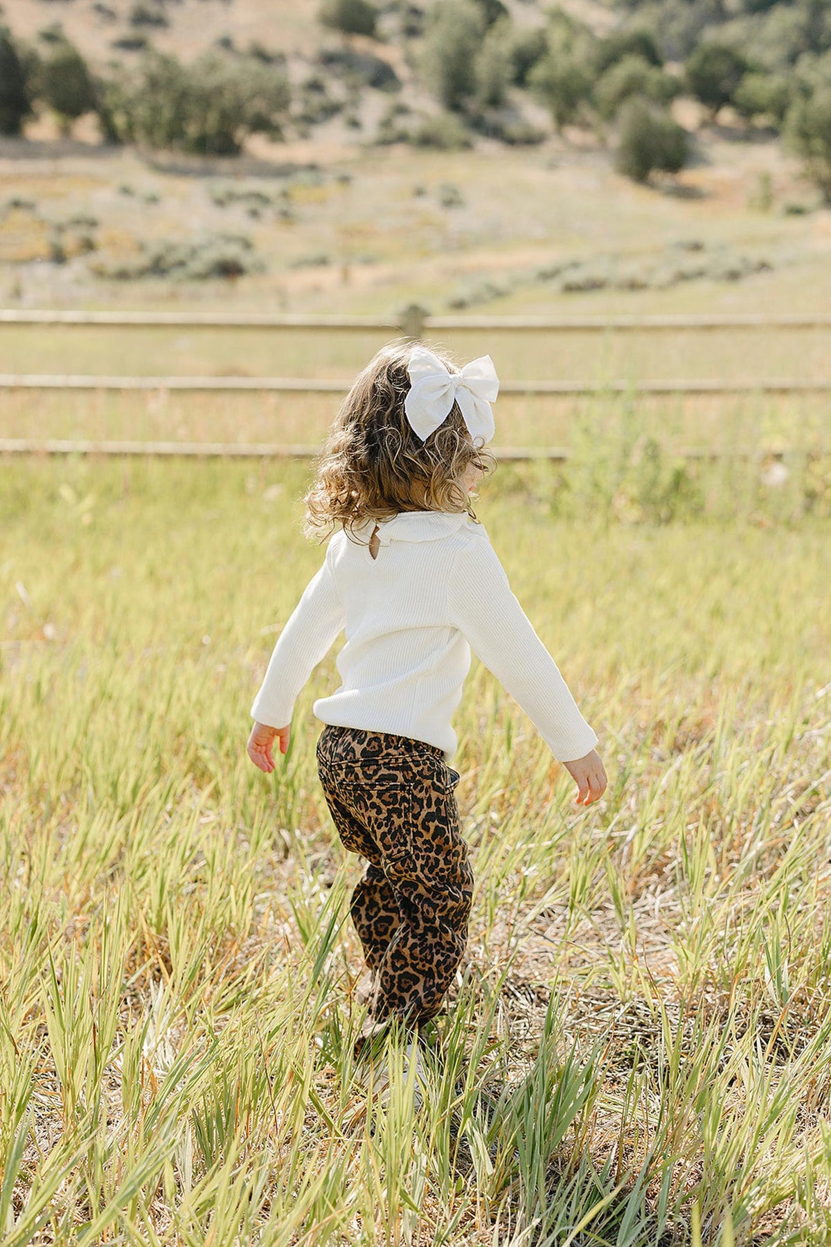 A young child walks through a grassy field wearing a white long sleeve top and leopard print pants, with a large white bow in their hair.