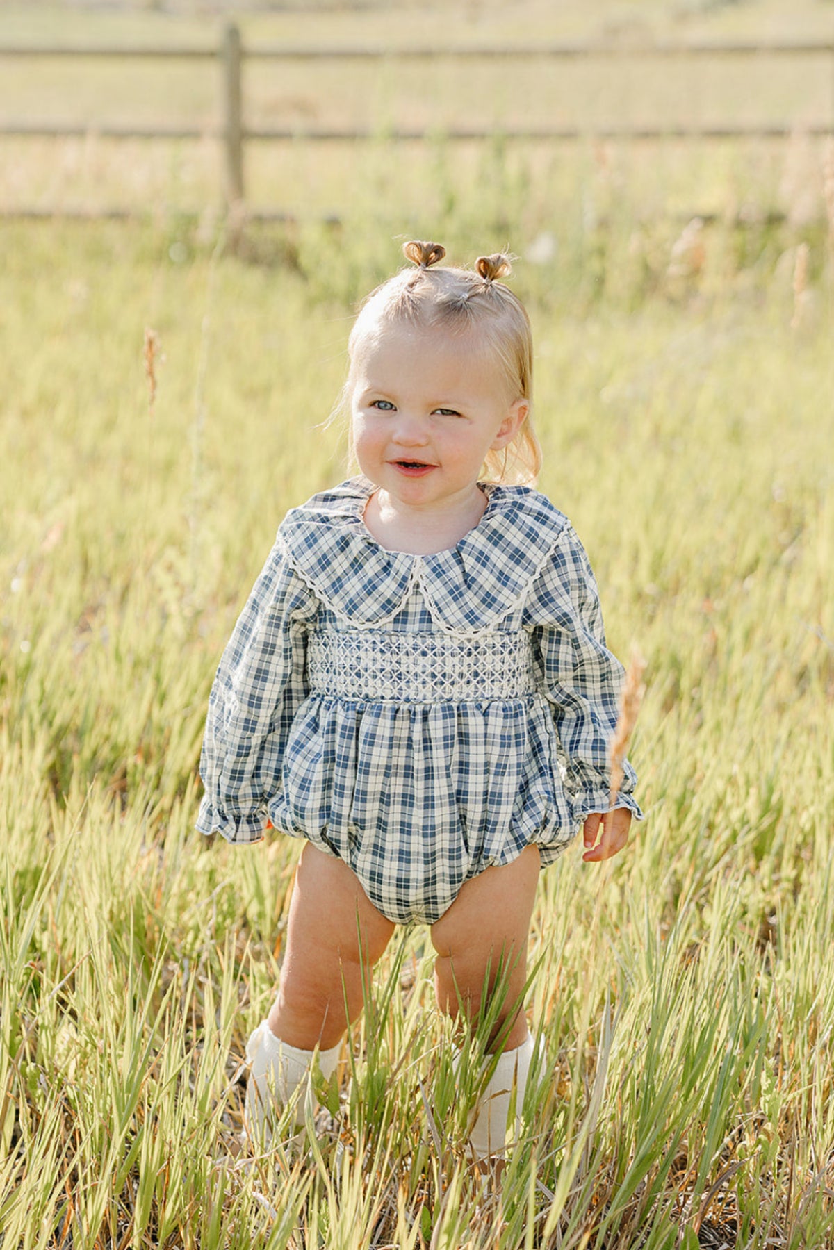 A smiling toddler stands in a grassy field, wearing a blue plaid romper with a ruffle collar. The outfit features long sleeves and is perfect for playful outdoor activities.