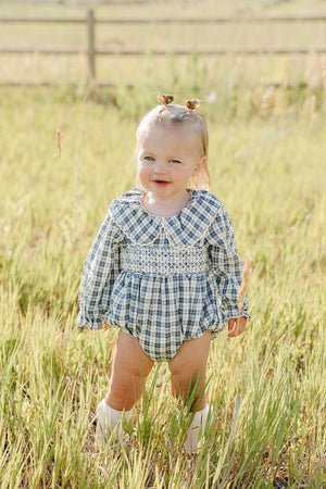 A smiling toddler stands in a grassy field, wearing a blue plaid romper with a ruffle collar. The outfit features long sleeves and is perfect for playful outdoor activities.