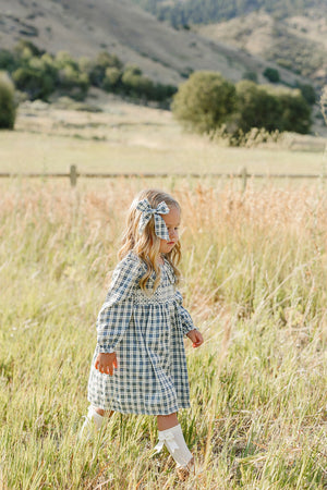 A young girl walks through a grassy field, wearing a blue plaid dress with a ruffle collar and long sleeves, complemented by a matching hair bow.