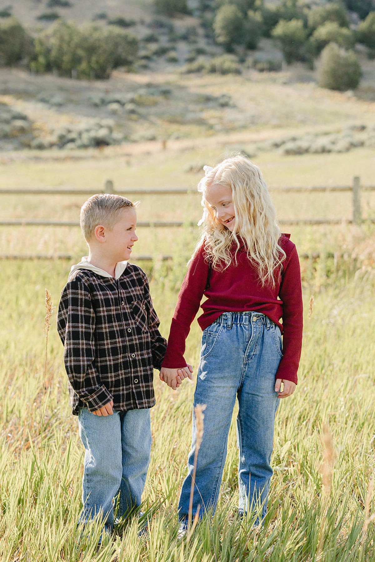 Two children stand in a grassy field, holding hands. One boy wears a black and plaid hoodie, while the girl is dressed in a burgundy long-sleeve top, both smiling happily.