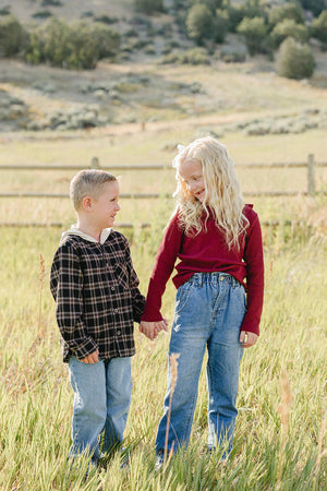 Two children stand in a grassy field, holding hands. One boy wears a black and plaid hoodie, while the girl is dressed in a burgundy long-sleeve top, both smiling happily.