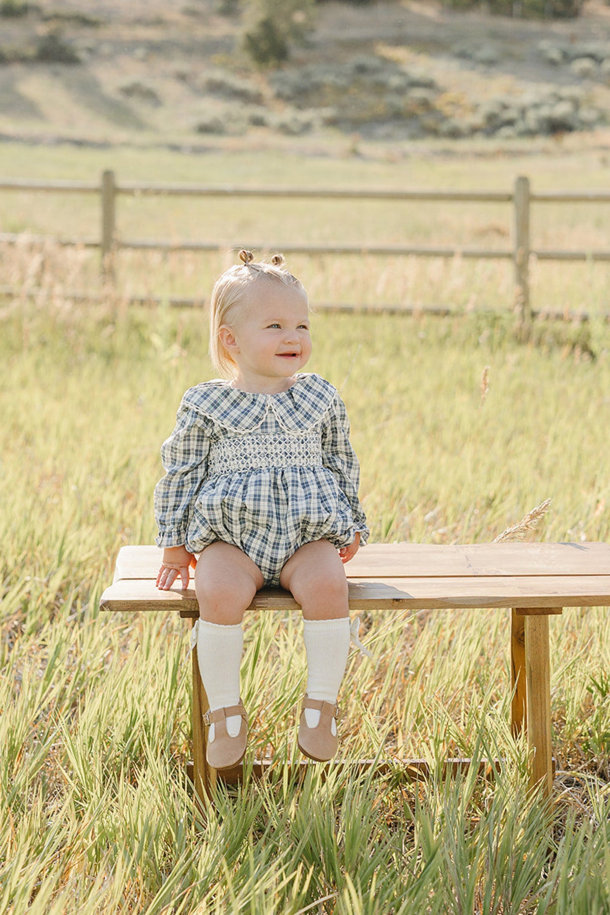 A smiling toddler sits on a wooden bench in a grassy field, wearing a blue plaid ruffle collar romper. The serene background features rolling hills and a wooden fence.