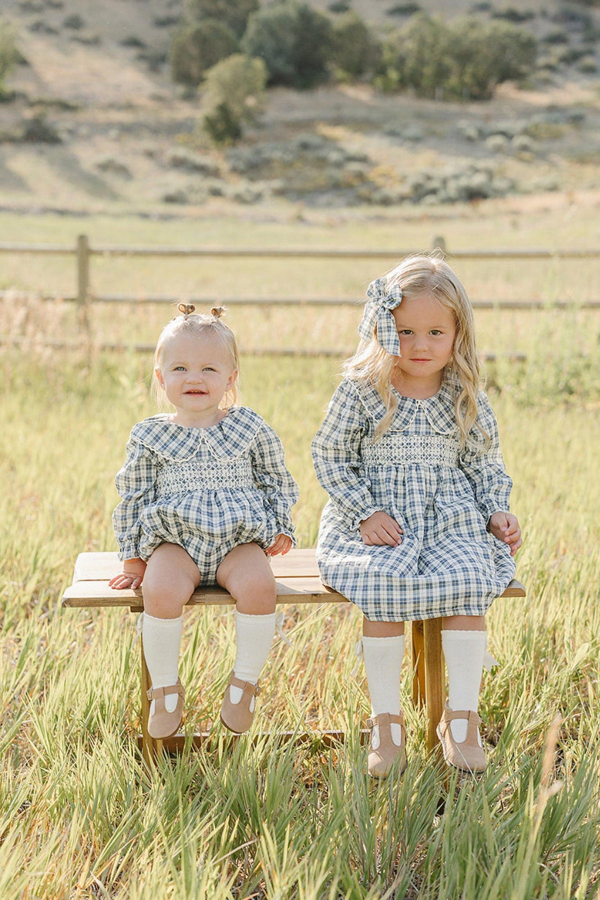 Two young girls sit on a wooden bench in a grassy field, wearing matching blue plaid dresses with ruffle collars. The setting features a serene, natural landscape in the background.