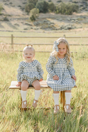 Two young girls sit on a wooden bench in a grassy field, wearing matching blue plaid dresses with ruffle collars. The setting features a serene, natural landscape in the background.
