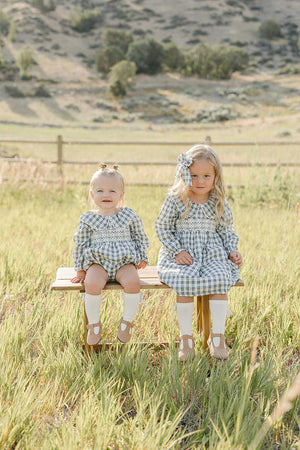 Two young girls sit on a wooden bench in a grassy field, wearing matching blue plaid outfits.