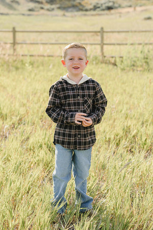 A young boy stands in a grassy field, wearing a dark plaid hooded button-up shirt and blue jeans.