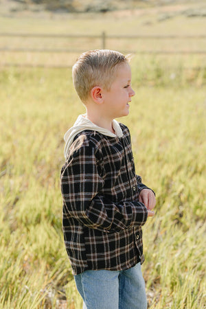 A young boy stands in a grassy field wearing a dark plaid hooded button-up shirt. The shirt features a classic plaid pattern and is paired with light blue jeans for.
