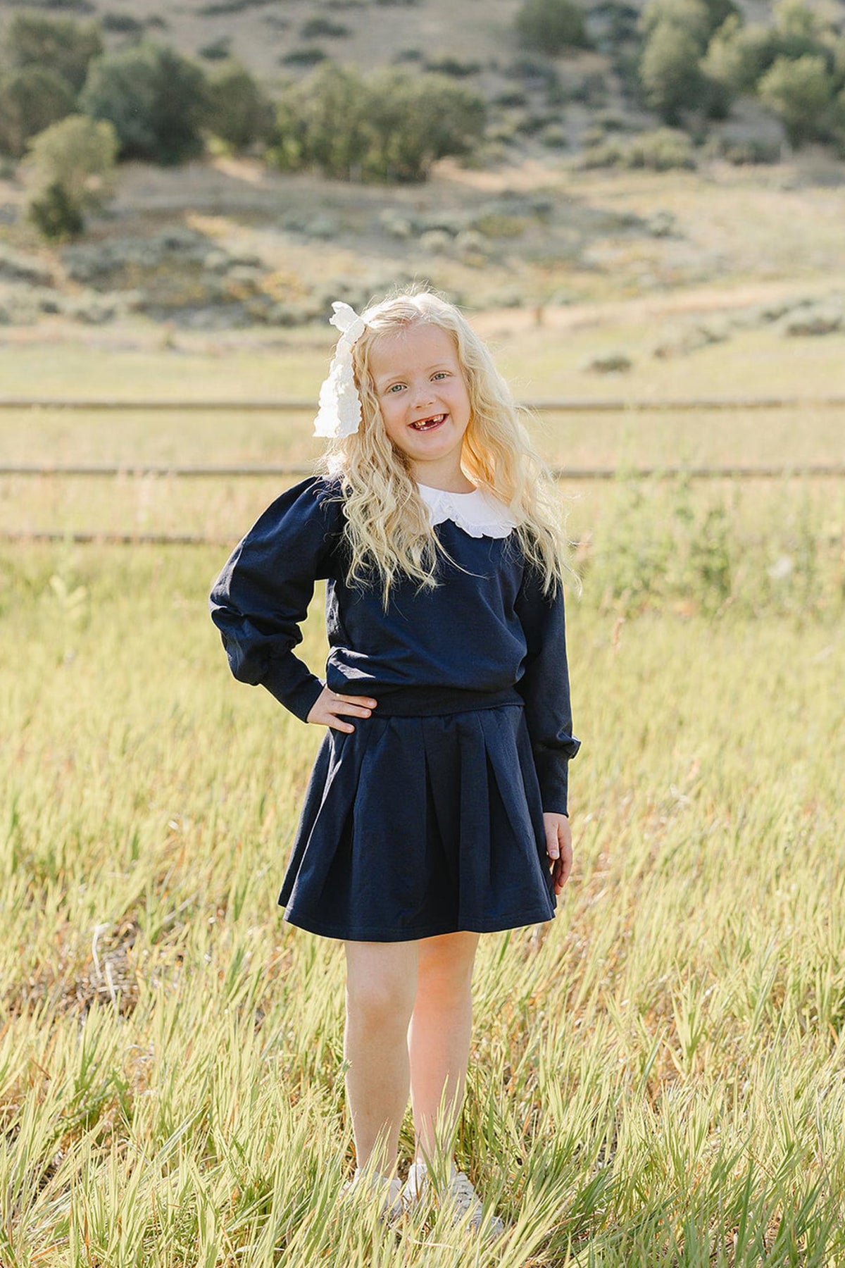 A young girl stands in a grassy field, wearing a navy blue pleated skirt set with a Peter Pan collar.
