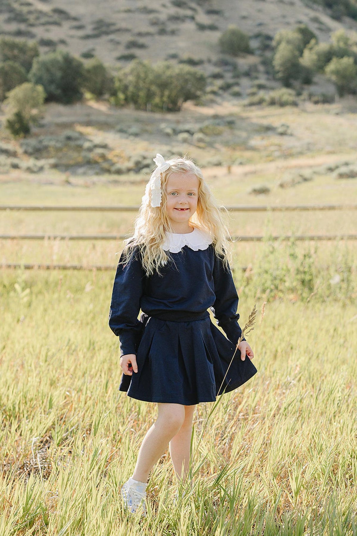 A young girl wearing a navy blue dress with a Peter Pan collar stands in a sunlit grassy field, surrounded by rolling hills and a wooden fence in the background.