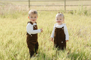 Two young children stand in a sunlit field, wearing brown corduroy overalls. The boy points while the girl smiles, both dressed in white long-sleeve tops.
