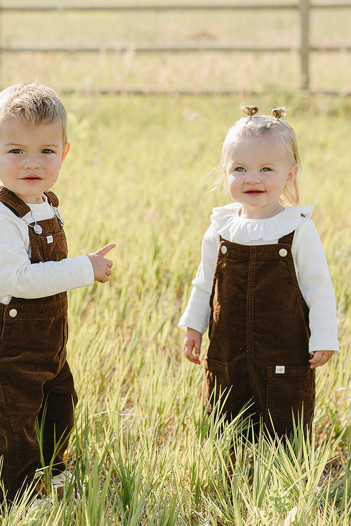 Two young children stand in a grassy field wearing brown corduroy overalls. The boy points playfully while the girl smiles, both dressed in cozy, casual outfits perfect for outdoor play.