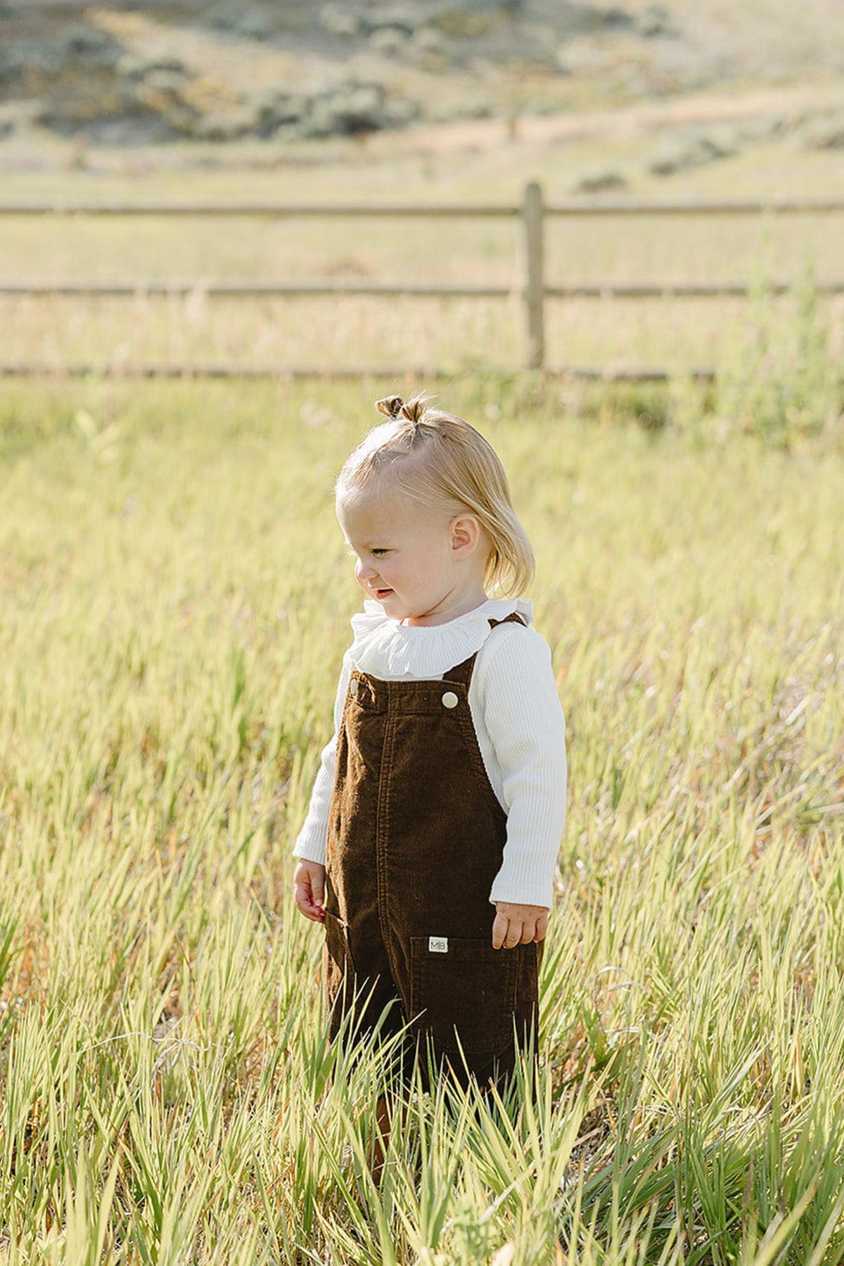 A young child stands in a grassy field wearing a brown corduroy dress over a white long-sleeve shirt.