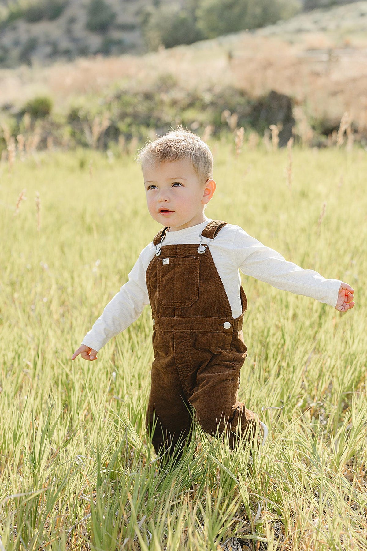 A young child wearing brown corduroy overalls and a white long-sleeve shirt stands in a grassy field, enjoying the outdoors. The setting features gentle hills in the background.