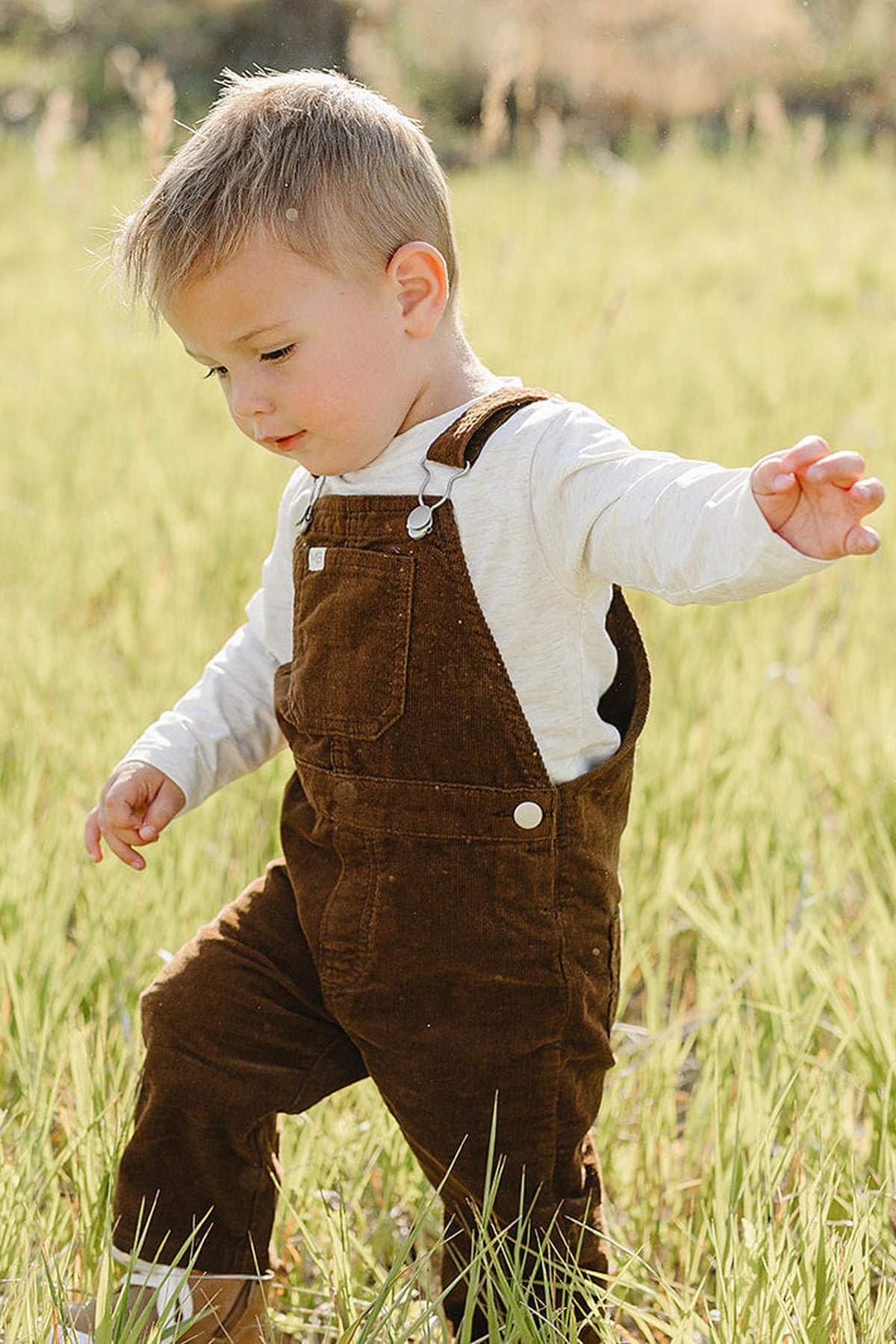 A young child dressed in brown corduroy overalls and a light long-sleeve shirt explores a grassy field, enjoying the outdoors with a playful expression. The setting is bright.