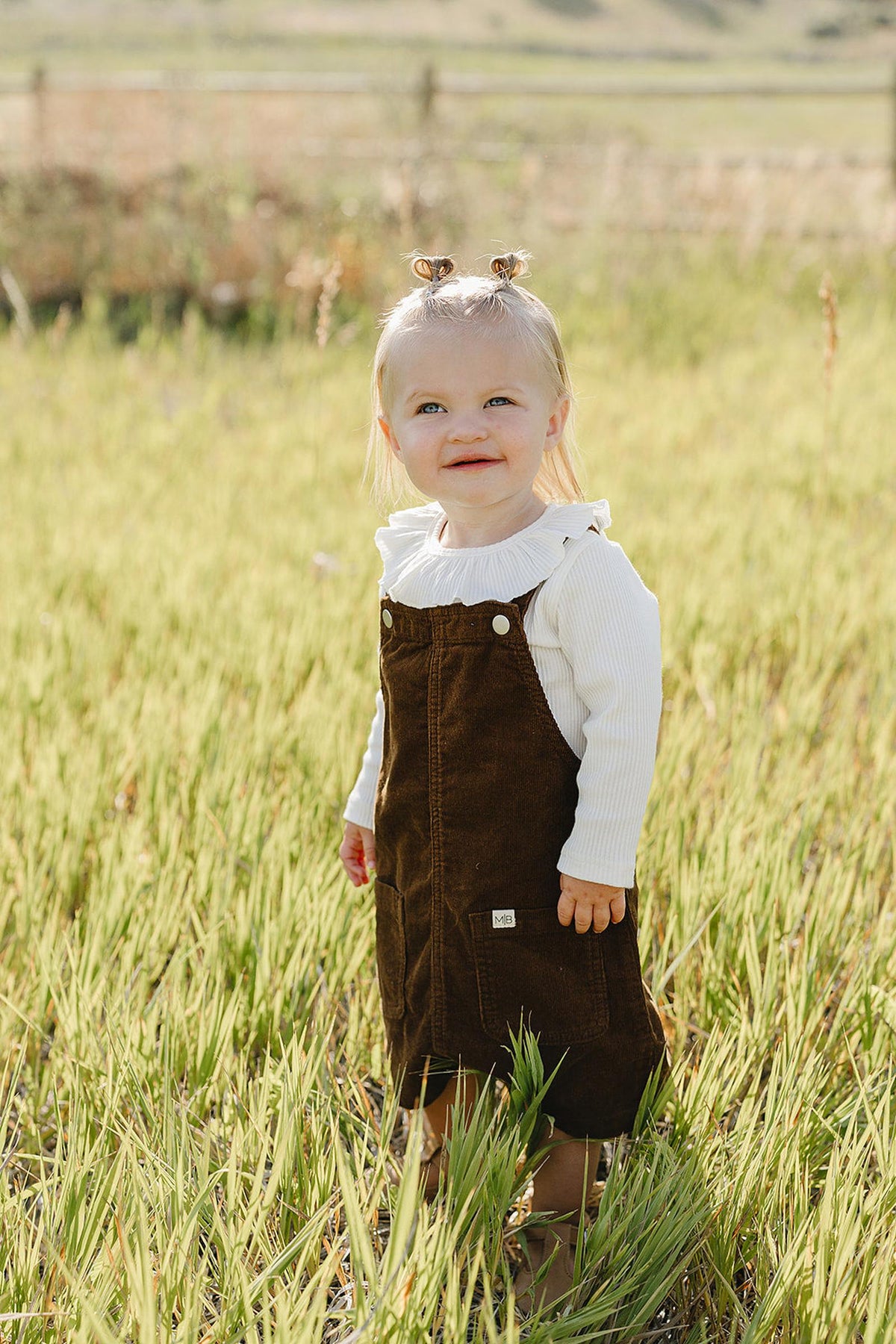 A smiling toddler stands in a grassy field, wearing a brown corduroy pinafore dress over a white long-sleeve shirt. The outfit is ideal for casual outings and seasonal layering.