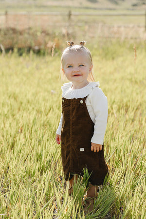 A smiling toddler stands in a grassy field, wearing a brown corduroy pinafore dress over a white long-sleeve shirt. The outfit is ideal for casual outings and seasonal layering.