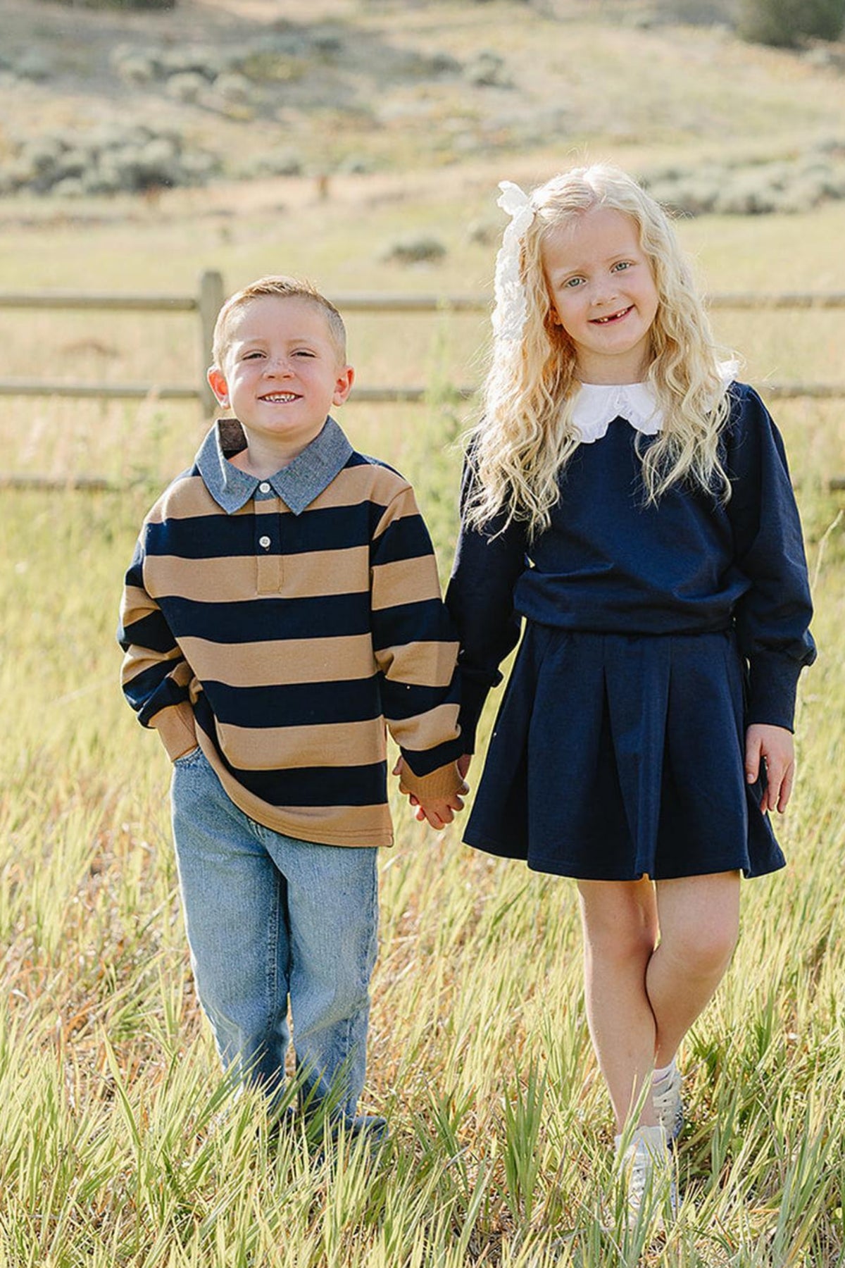 Two children stand hand in hand in a grassy field, wearing a striped navy and camel shirt and a solid navy dress, surrounded by a serene natural landscape.