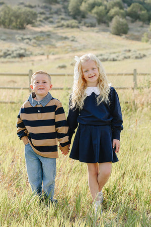 A boy in a striped navy and tan sweater and a girl in a navy pleated skirt set walk hand-in-hand through a grassy field, surrounded by trees and hills.