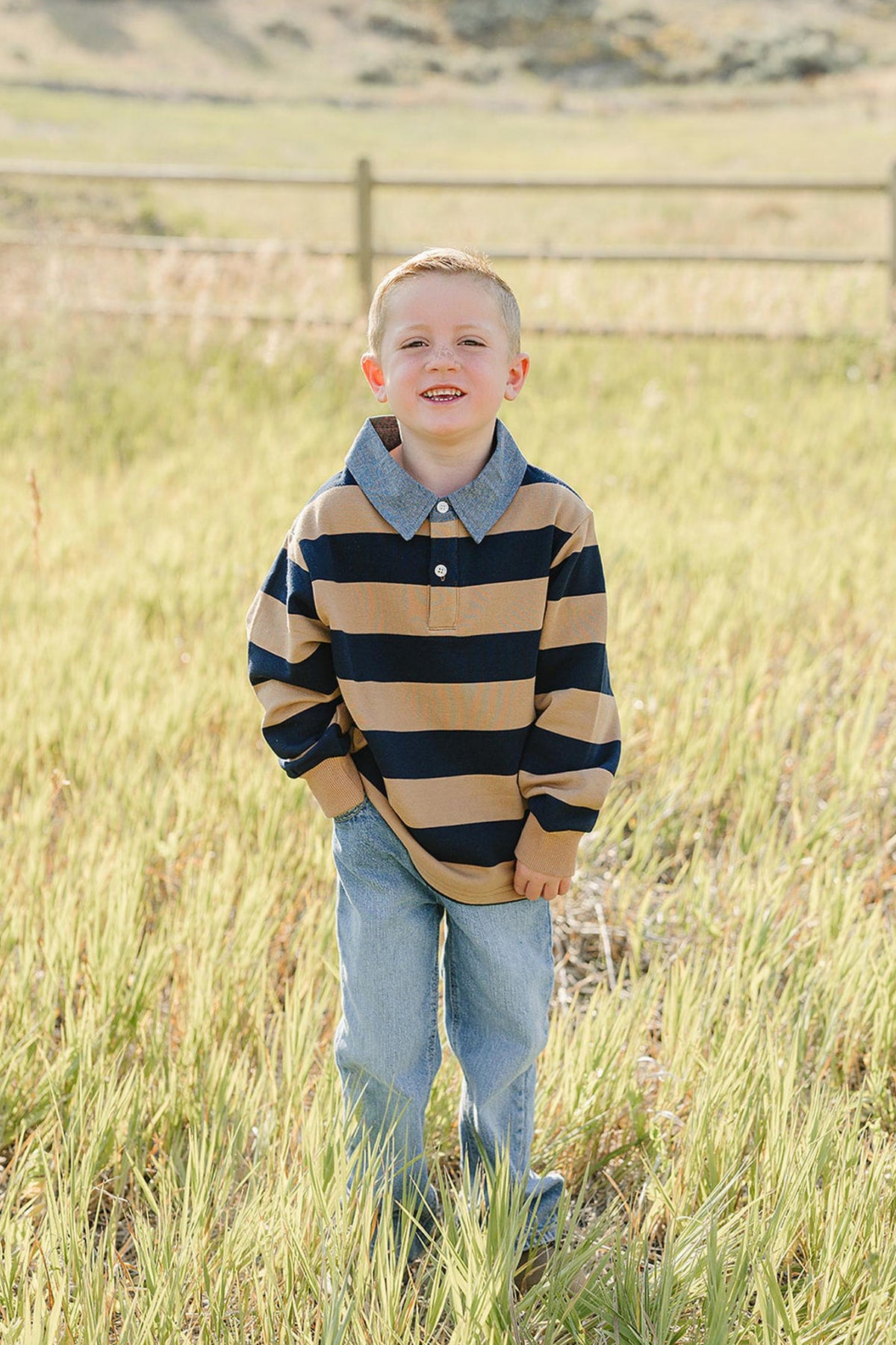 A young boy smiles while standing in a field of tall grass, wearing a classic camel and navy striped collar shirt with a denim collar, paired with blue jeans.