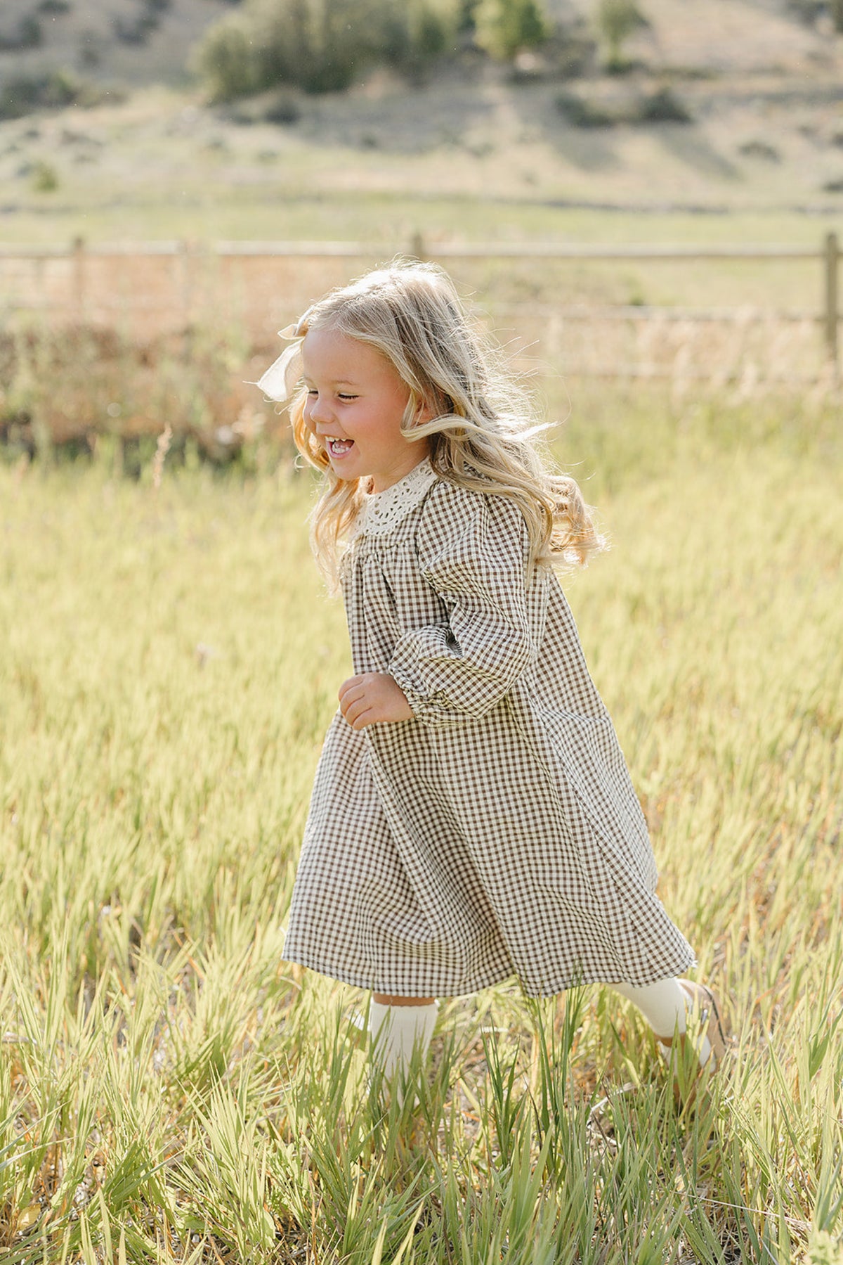 A joyful little girl runs through a field, wearing a camel gingham dress with a lace collar. The dress features soft cotton material, perfect for playful autumn days.