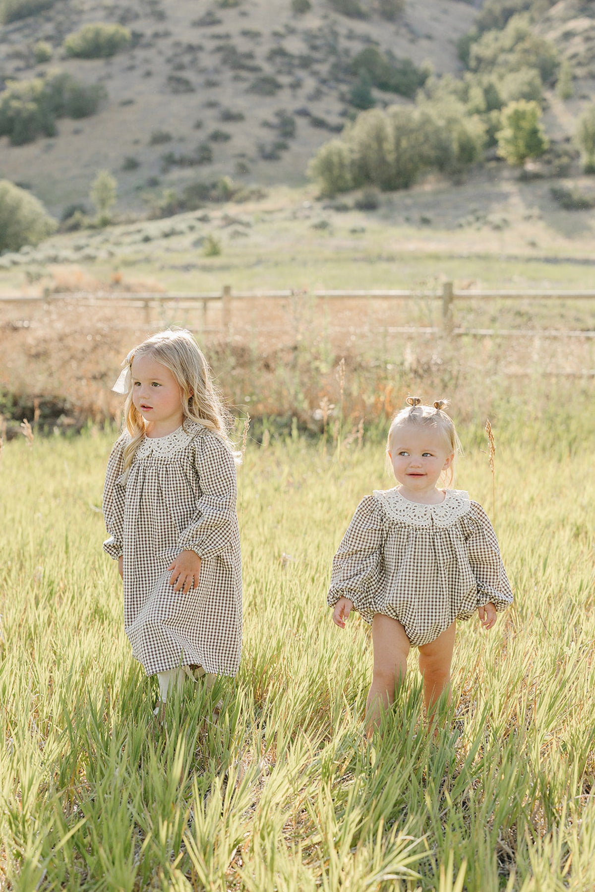 Two young girls play in a sunlit field, wearing gingham-patterned dresses. One girl is in a long-sleeve dress, while the other wears a short romper with a lace collar.