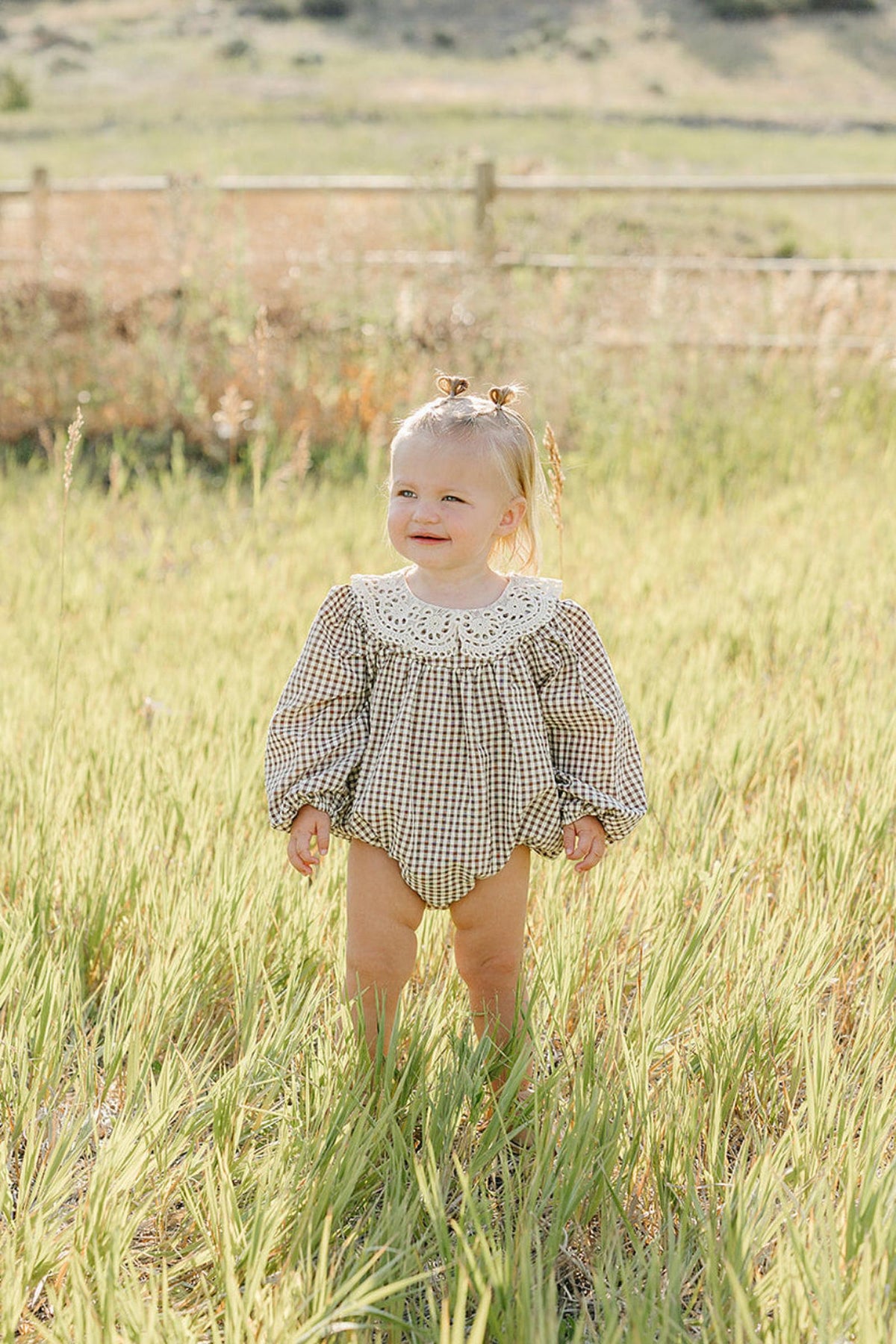 A toddler stands in a grassy field wearing a camel gingham lace bubble romper with a white collar. The soft cotton fabric creates a comfortable look perfect for outdoor play.